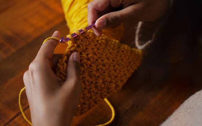 close-up of a lady's hand crocheting using an aluminium crochet hook needle on a yellow yarn
