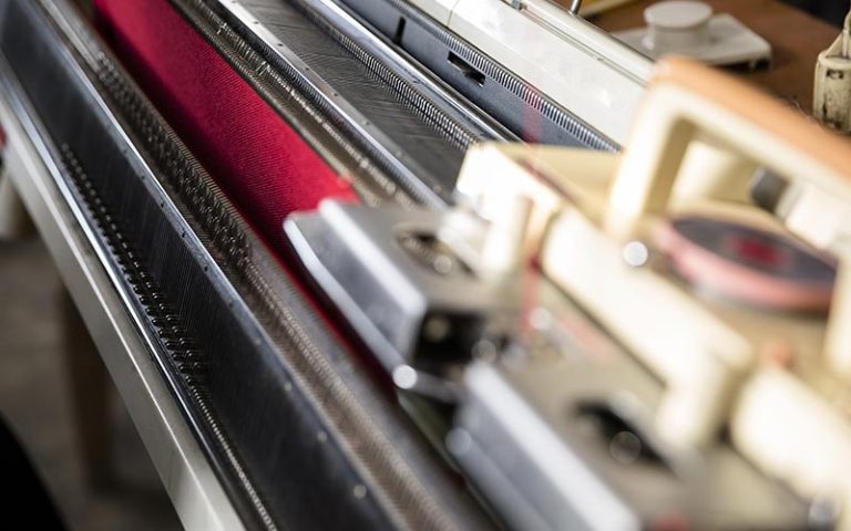 Close-up of a flatbed machine with a red yarn being knitted into a dress