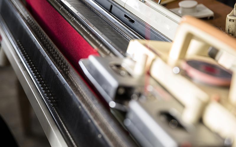 Close-up of a flatbed machine with a red yarn being knitted into a dress